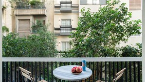 A balcony with two wooden chairs, a round table with a fruit bowl and a can, and a view of a building with balconies and greenery. - A short walk to Syntagma and Plaka (Athens)