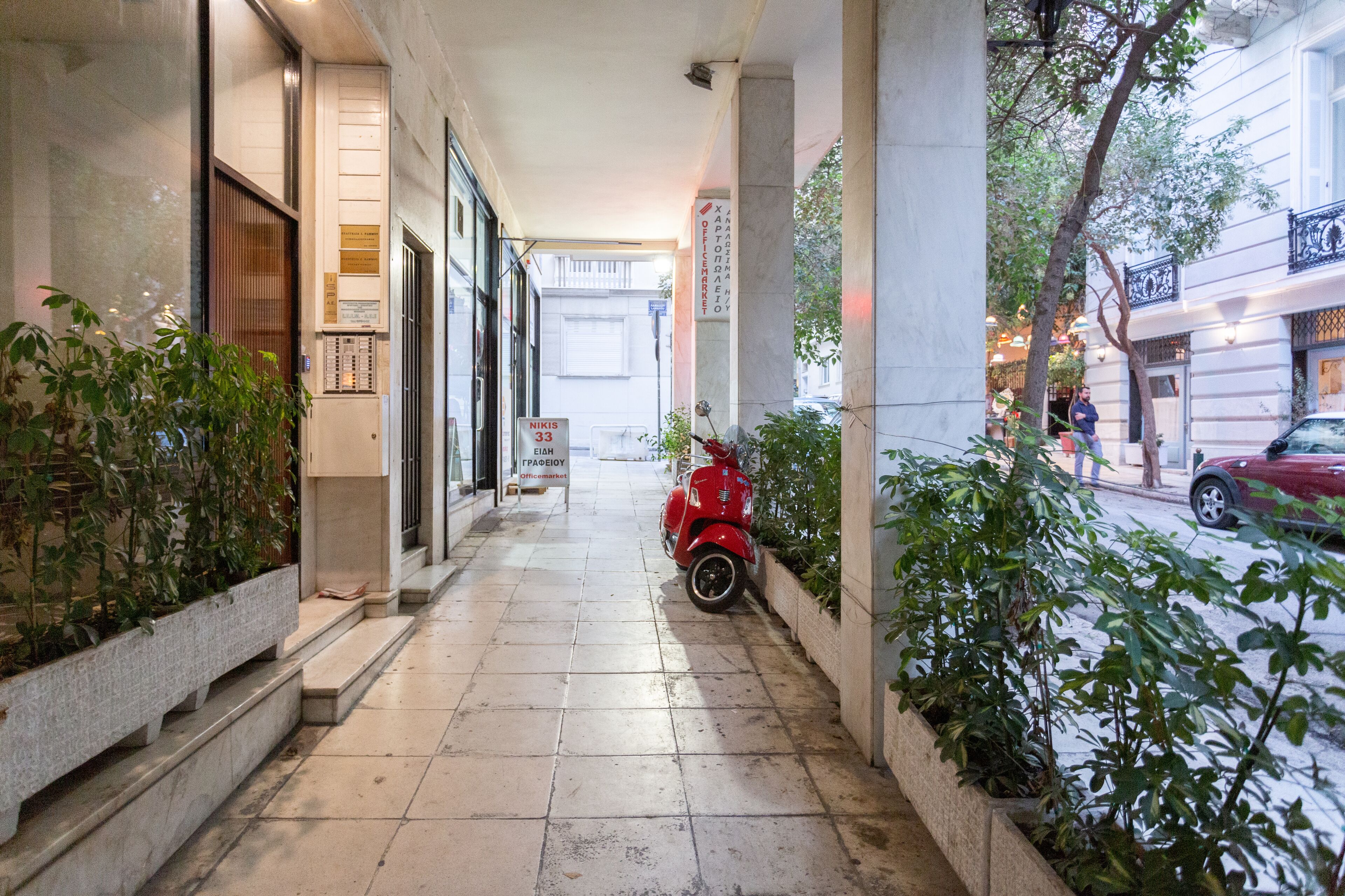 A corridor with a red scooter parked on the right, potted plants on both sides, and a sign with text in a foreign language.