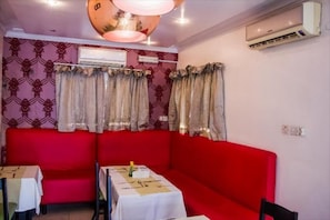 A dining area with red cushioned seating, tables with white tablecloths, patterned wallpaper, and an air conditioning unit on the wall. - Nne-Era Residence (Lagos)