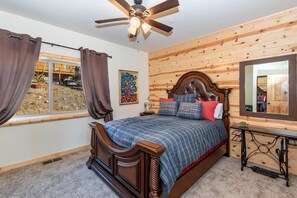 A bedroom with a wooden headboard, a ceiling fan, a large window with curtains, and a mirror on the wall.