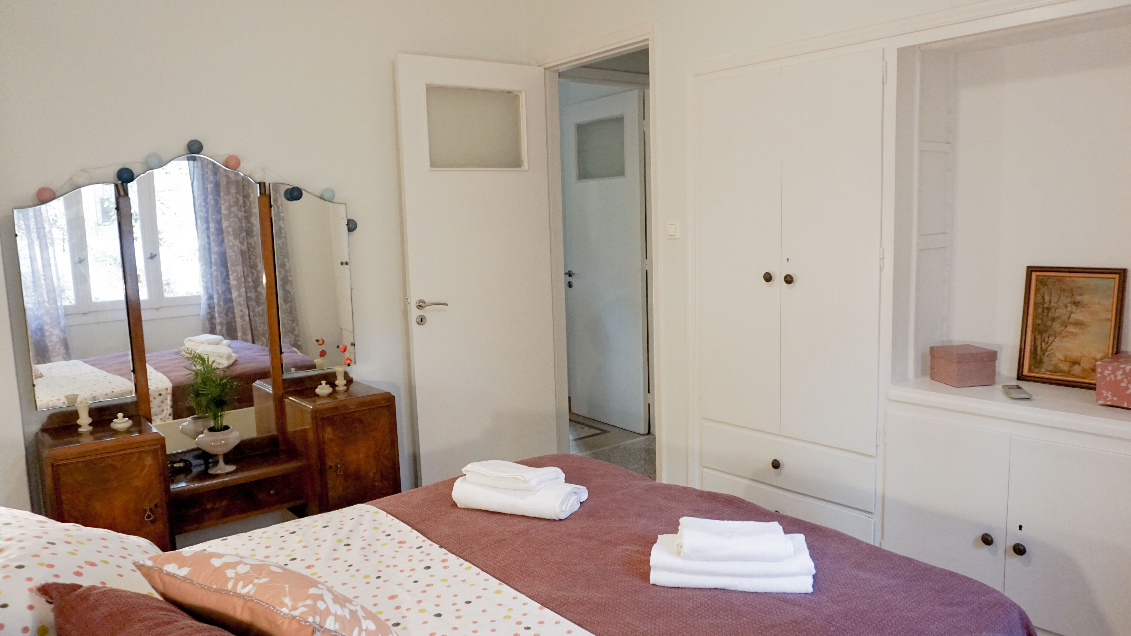 A bedroom with a bed, a dresser, a mirror, a door leading to another room, and a small cabinet with a picture.