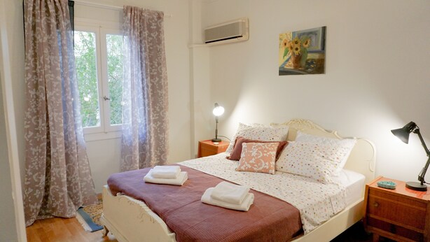 Een slaapkamer met een bed, een nachtkastje met een lamp, een raam met gordijnen en een schilderij aan de muur.