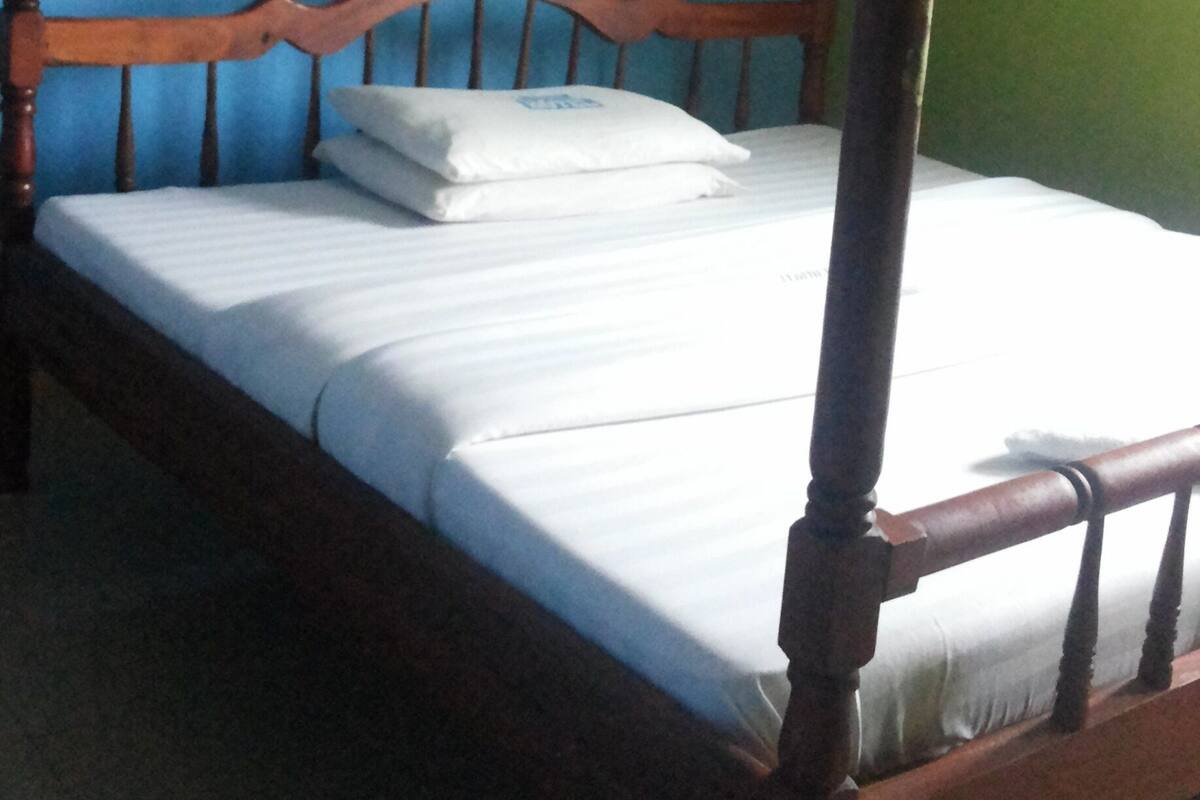 A single bed with a wooden frame and white bedding, a bedside table, and a wall-mounted fan.