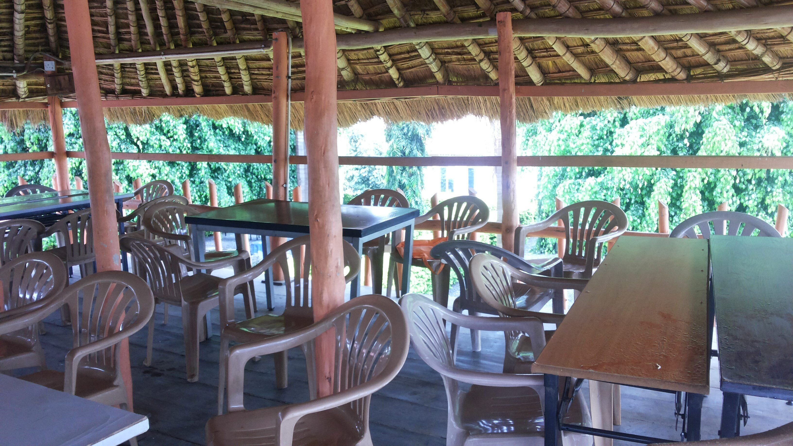 An outdoor dining area with plastic chairs and tables, a thatched roof, and greenery in the background.