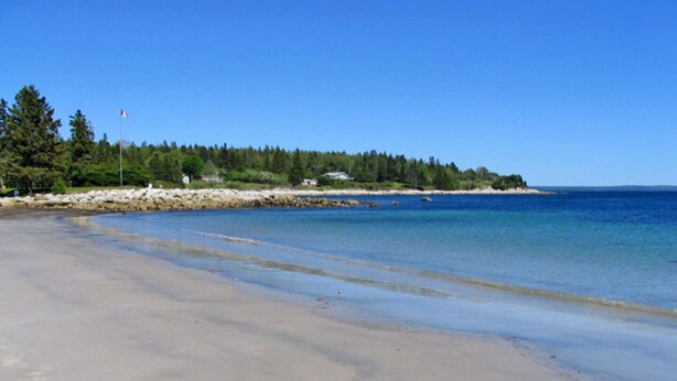 A beach with clear blue water, sandy shore, and a forested area with a flag in the background. - Anchorage House & Cottages - Bayswater Cottage (Hubbards)