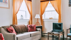 A living room with a sofa, a television on a stand, a lamp, and a window with curtains. - Anchorage House & Cottages - Bayswater Cottage (Hubbards)