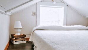 A bedroom with a sloped ceiling, a bedside lamp, a bed with a white cover, and a window with a view. - Anchorage House & Cottages - Bayswater Cottage (Hubbards)