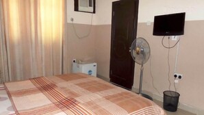 Una habitación con una cama, un ventilador de pie, un televisor montado en la pared y un pequeño calentador.