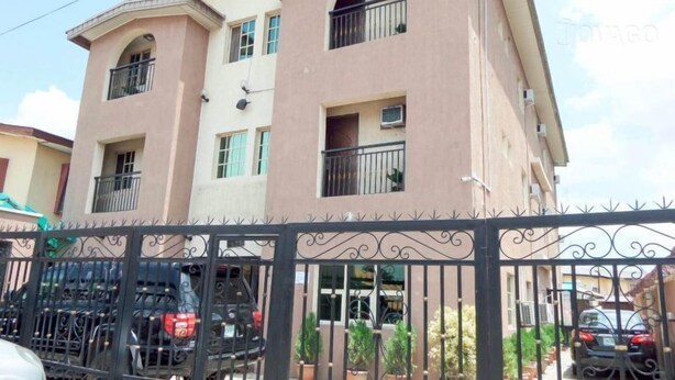 Un edificio residencial de varios pisos con balcones, entrada protegida por un portón y automóviles estacionados.