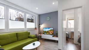 Ett modernt vardagsrum med en grön soffa, ett litet vitt bord, en platt-TV och en krukväxt.