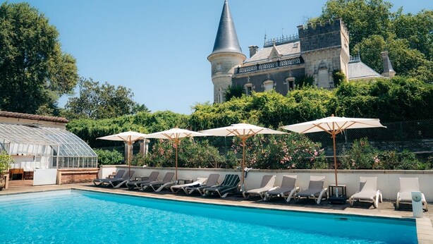 Ein Poolbereich mit Liegestühlen und Sonnenschirmen, ein Gewächshaus und im Hintergrund ein schlossartiges Gebäude.