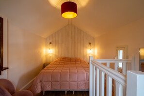 A bedroom with a bed, a wooden sloped ceiling, two wall-mounted lights, and a white railing.