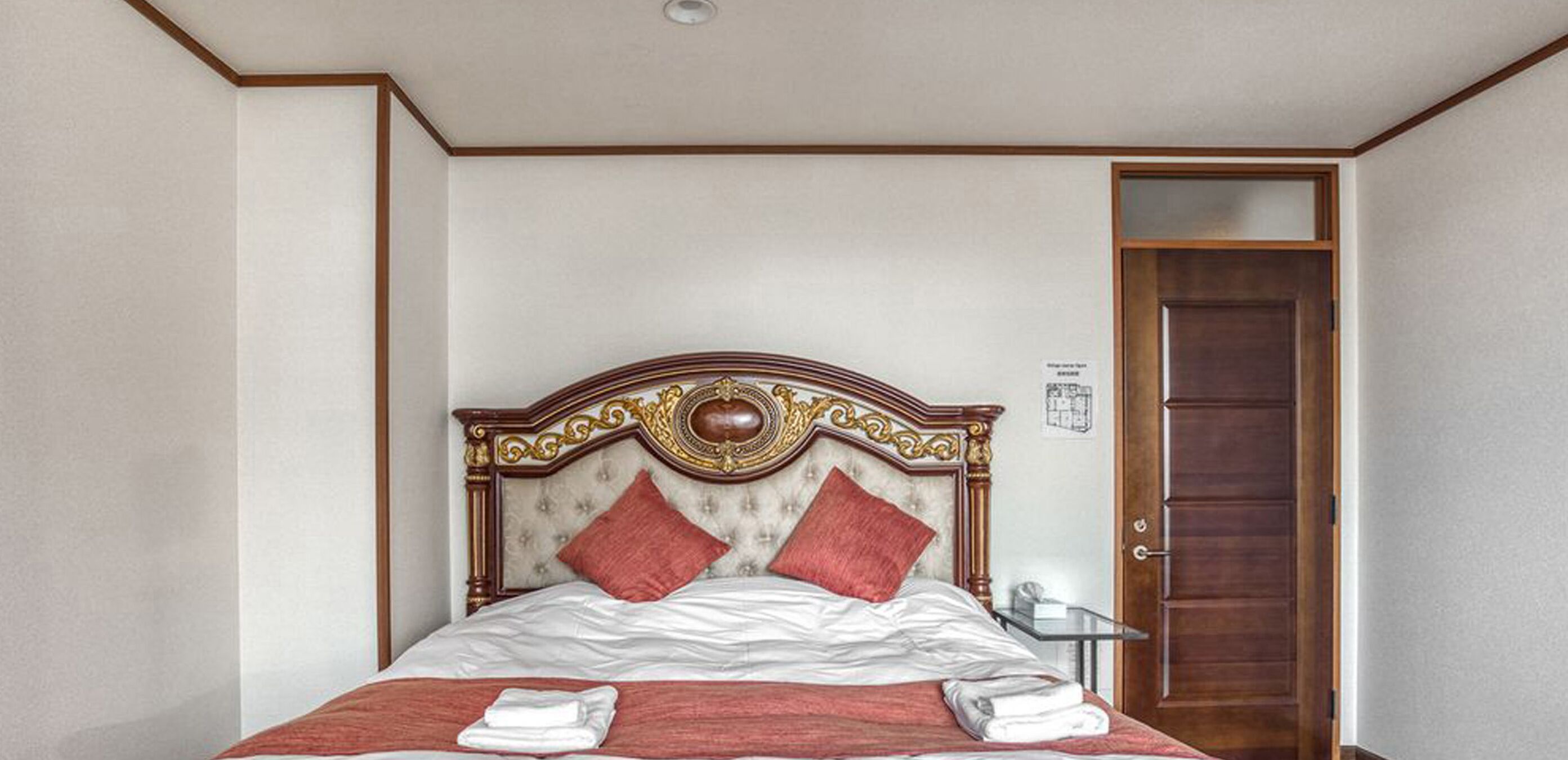 A neatly made bed with a wooden headboard, two red pillows, and a folded white towel on the bed.