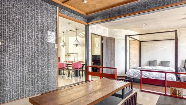 A modern hotel room with a large bed, a dining area with a wooden table and chairs, and a view through a glass door to a dining area with red chairs. - Woo House (Osaka)