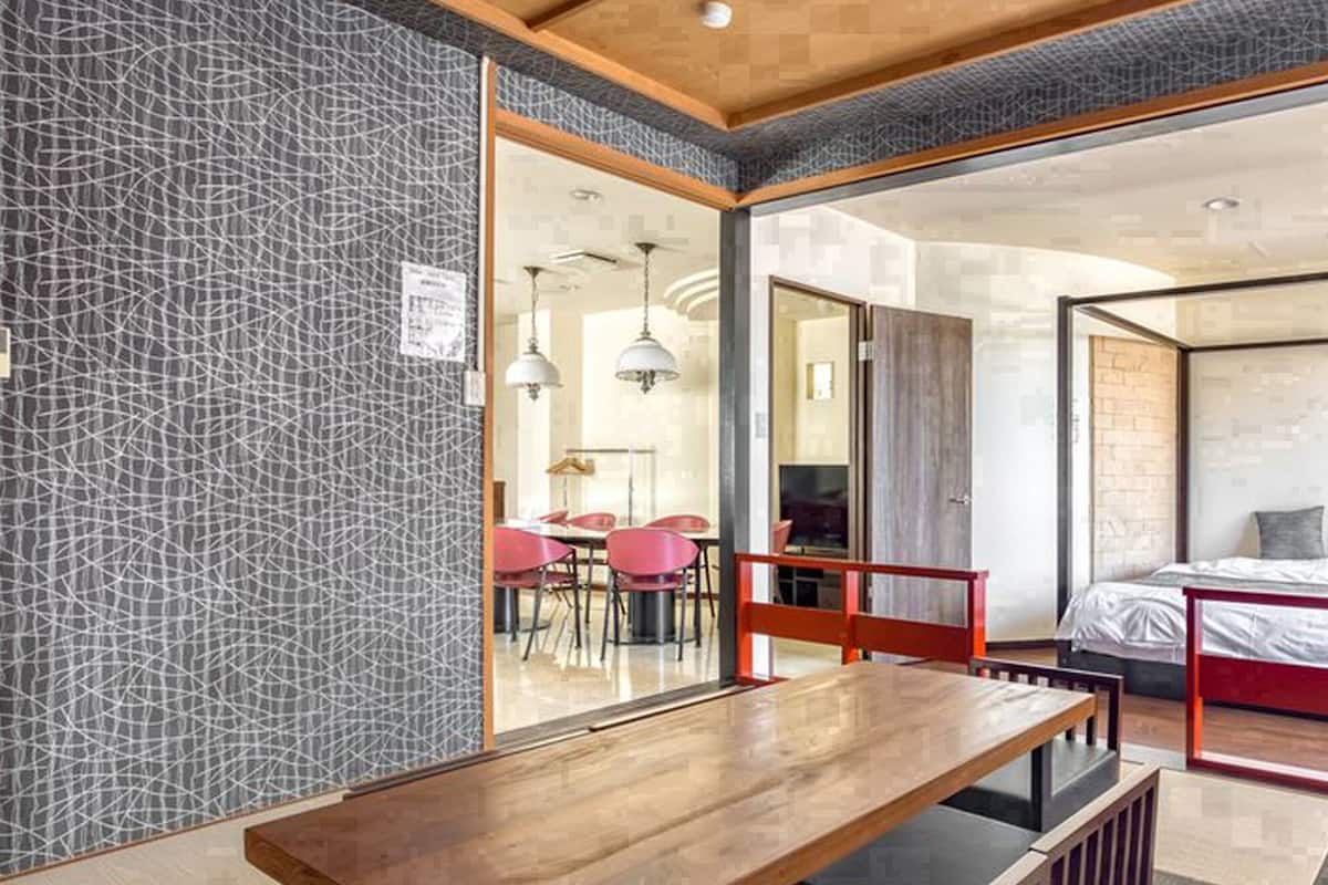Une chambre d’hôtel moderne dotée d’un grand lit, d’un coin repas avec une table et des chaises en bois, et offrant une vue sur un espace de restauration avec des chaises rouges, visible à travers une porte vitrée.
