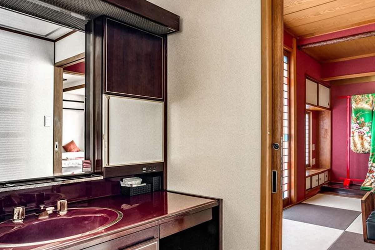 Une salle de bain avec un grand miroir, un meuble-lavabo et une vue sur une pièce dont la fenêtre est ornée d’un rideau vert.