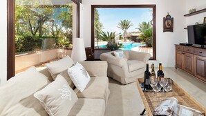 Ein Wohnzimmer mit einer Couch, Sesseln, einem Couchtisch mit Weinflaschen und Gläsern, einem Fernseher und Blick auf einen Swimmingpool und Grünflächen.