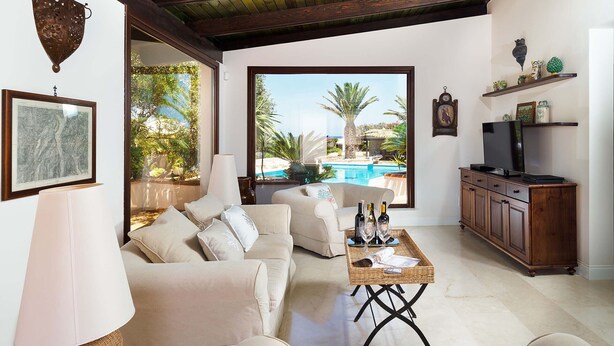 Ein Wohnzimmer mit einem beigefarbenen Sofa, einem Couchtisch mit Flaschen, einem Fernseher auf einer Holzkommode und einem großen Fenster mit Blick auf einen Pool.