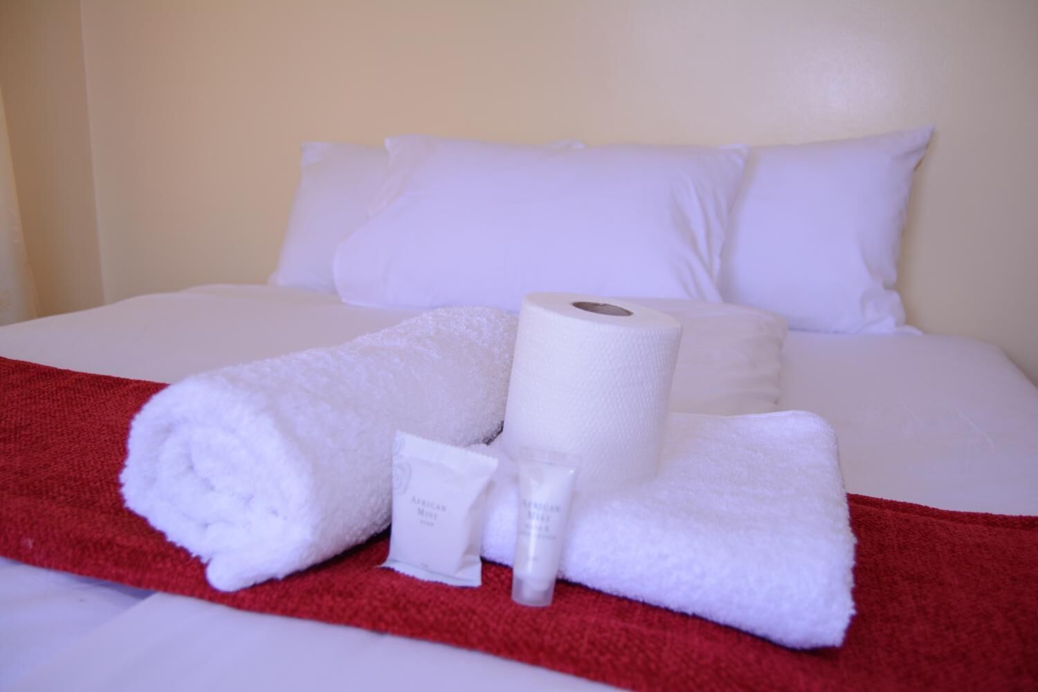 Una cama bien hecha con ropa blanca, una manta roja y artículos de aseo, incluyendo una botella de loción y un tubo de crema.