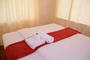 A neatly made bed with white linens and a red throw blanket, a roll of toilet paper, and a folded white towel on the bed. - Royal Guest House (Harare)