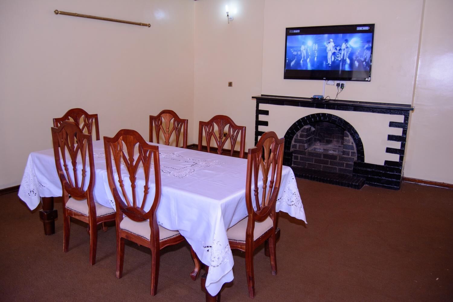 Un comedor con una larga mesa, seis sillas de madera, un mantel blanco, un televisor en la pared y una chimenea.