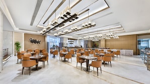 A spacious dining area with modern lighting fixtures, multiple tables and chairs, and a view of the kitchen in the background. - Kare Hotels (Shenzhen)