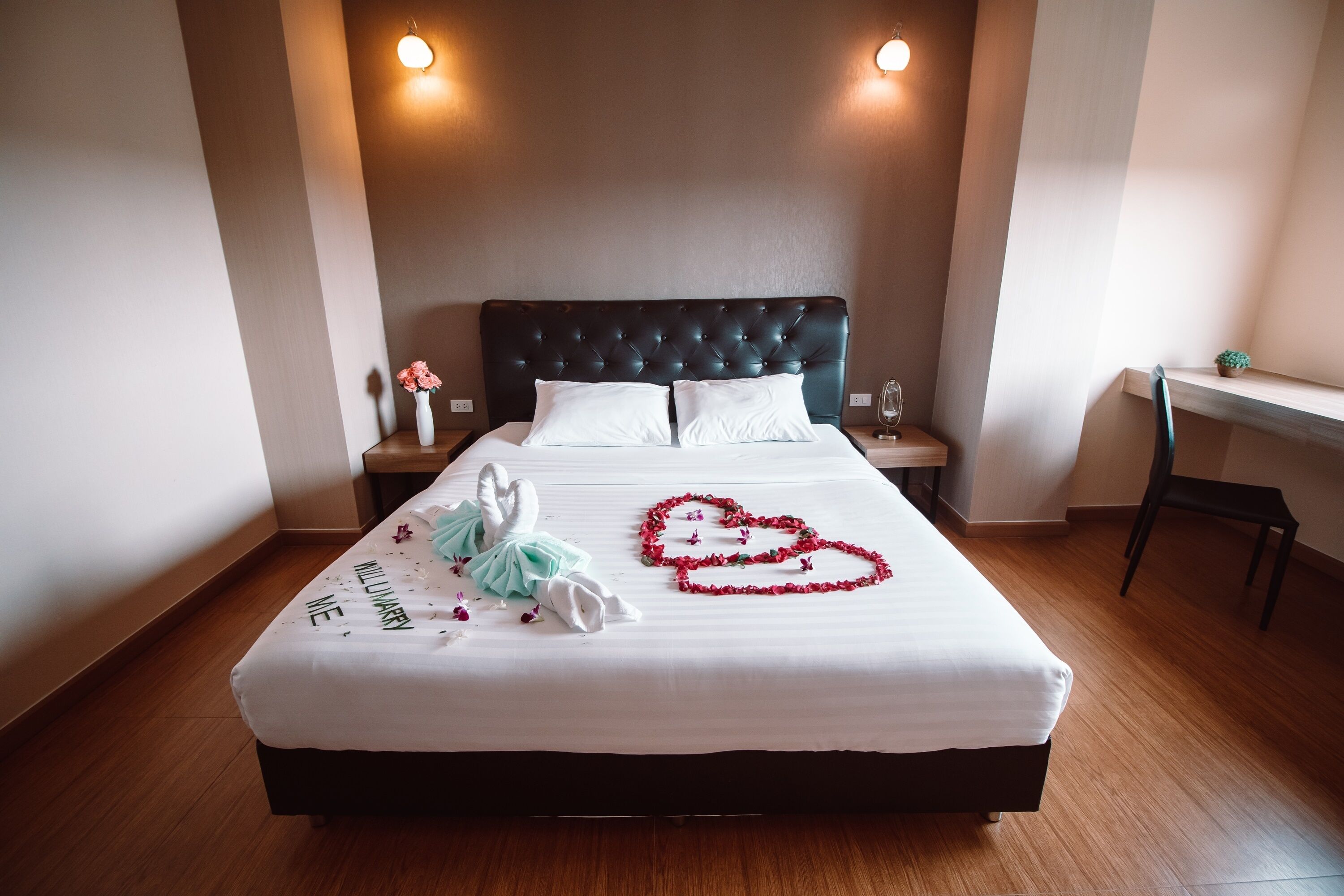 Una cama bien tendida con sábanas blancas, cabecera acolchada y un arreglo floral con forma de corazón sobre el edredón.
