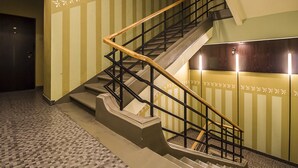 Un escalier avec une main courante en bois et des rampes en métal noir, menant à un palier doté d’un mur à motifs et d’une porte visible au fond.
