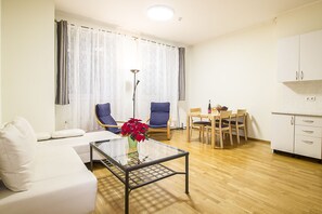 A modern living room with a white sofa, a glass coffee table with a vase of red flowers, two blue chairs, and a dining area with a wooden table and chairs.
