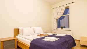 Un lit bien fait, recouvert d’une couverture violette, une table de chevet en bois, un petit plateau avec des croissants et une fenêtre avec des rideaux.