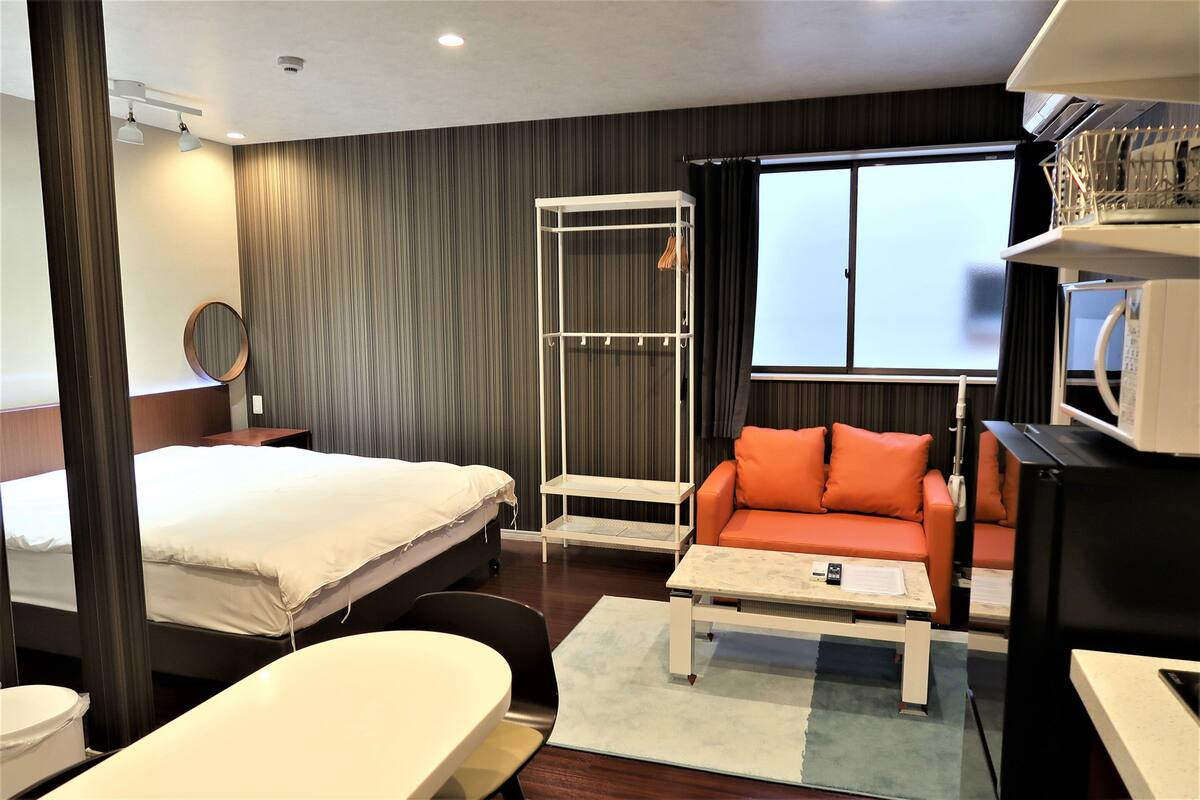 Quarto de hotel com cama, sofá, mesinha, espelho, guarda-roupa e janela.