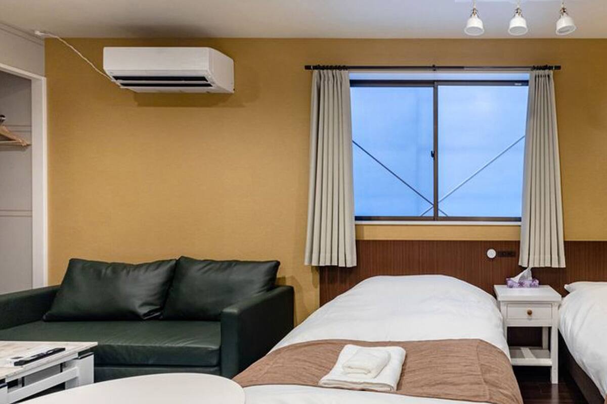 Quarto de hotel com duas camas, um sofá, uma mesinha de cabeceira, uma luminária e uma janela com cortinas.