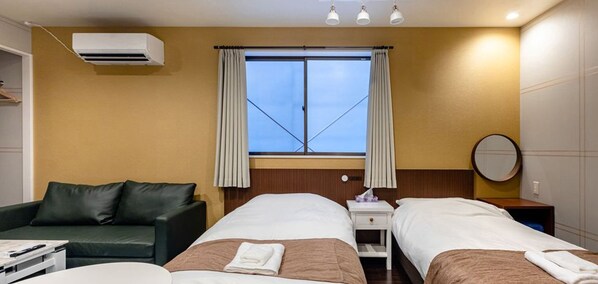A hotel room with two beds, a sofa, a bedside table, a lamp, and a window with curtains.