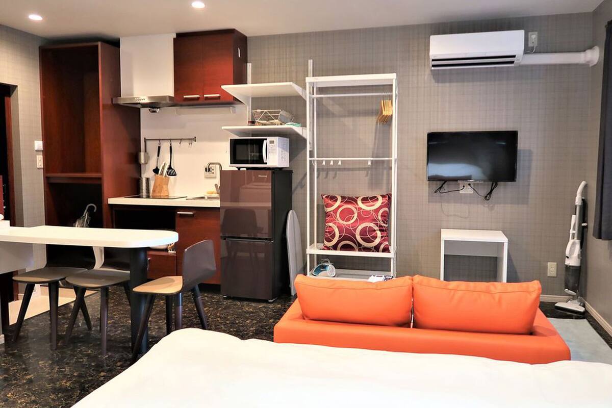 Quarto de hotel compacto com cozinha compacta, uma cama com almofadas laranja, TV e ar condicionado.