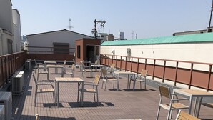 Una terraza al aire libre con mesas y sillas, un edificio al fondo y una estructura con techo verde.