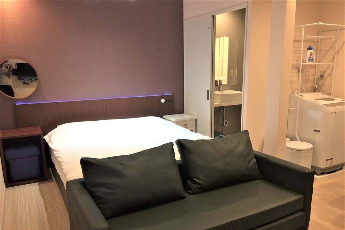 Quarto de hotel moderno com cama, sofá escuro, mesa de cabeceira, espelho e banheiro com vaso sanitário, pia e chuveiro.