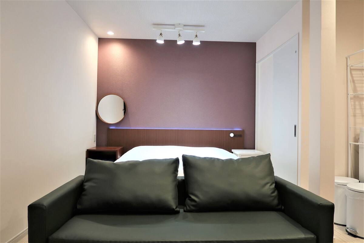 Quarto de hotel moderno com uma cama, um sofá de cor escura, um espelho redondo e um candeeiro no teto.