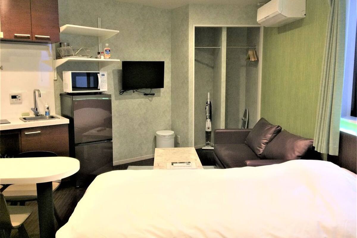Quarto de hotel com cama, escrivaninha e cadeira, micro-ondas, televisão, chuveiro e janela com cortinas.