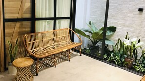 Un banco de mimbre con un taburete redondo, un colgante de luz y vistas a una pared de ladrillo blanco y plantas a través de amplios ventanales.