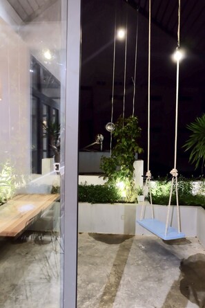 Un espacio al aire libre con una zona de estar, un columpio y una mesa, rodeado de vegetación y un edificio al fondo.