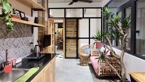 Cocina compacta con lavamanos, hervidor y diversos utensilios. Hay una silla de mimbre y una mesita con una planta. Una puerta corredera da acceso a un baño con ducha e inodoro.