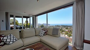 A modern living room with a beige sectional sofa, a wooden coffee table, and a large window offering a view of a coastal landscape. - Luxury villa with spectacular sea views, Pvt Pool, Gardens and Parking (Teulada)