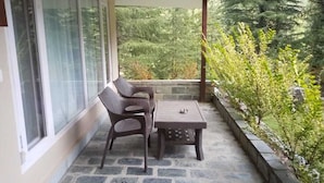 A covered patio with a stone floor, two wicker chairs, a small table, and a view of a forested area. - Asaka Himalayan Inspiration lodge Kasol (Bhuntar)