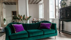 Ein grünes Samtsofa mit violetten Kissen in einem Raum, der ein großes Spiegel, einen Schrank und ein Fenster beherbergt.