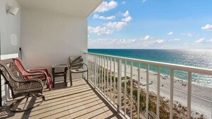 Ein Balkon mit Korbstühlen und einem kleinen Tisch mit Blick auf einen Strand, Menschen und einen klaren Himmel.
