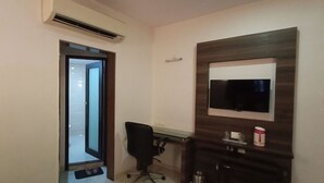 A hotel room with a desk, chair, TV mounted on a wooden panel, and an air conditioning unit. - Hotel Prime (Ahmedabad)