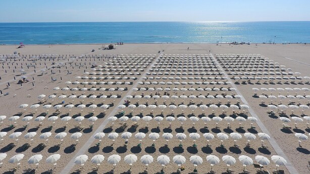 Ein Strand mit Reihen von Sonnenschirmen und Liegestühlen, einem klaren Himmel und dem Meer im Hintergrund.