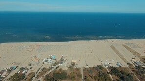 Ovenudblik over en strand med mange parasoller og liggestole, et parkeringsområde og et par bygninger.
