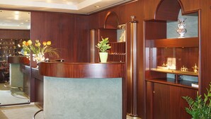 A reception desk with a potted plant, wooden paneling, and a display cabinet with decorative items. - As-Salam Aparthotel (Friedrichsdorf)