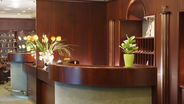 A reception desk with a wooden finish, a green potted plant, and a vase with flowers. - As-Salam Aparthotel (Friedrichsdorf)
